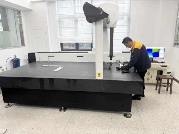En person står vid en stor svart mätbänk i ett modernt laboratorium. Mätbänken har en grå yta med några kontroller och verktyg på den. Ovanför bänken finns en mätarm kopplad till en datorskärm som visar färgade grafer. Rummet har ljusa väggar och stora fönster som släpper in naturligt ljus. En liten stol står i närheten.