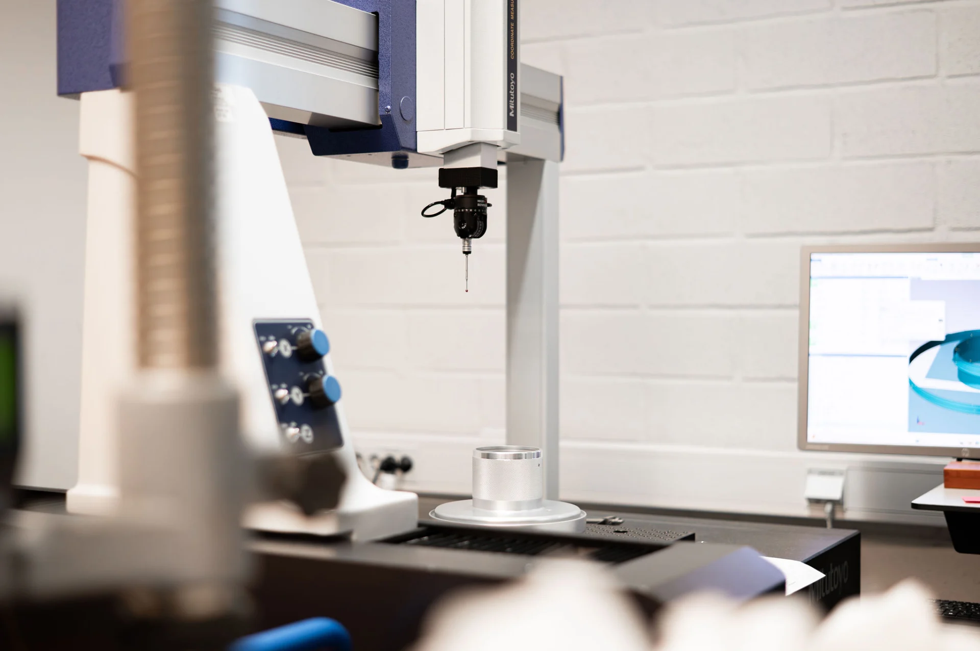 A precision measuring machine is in focus, with a probe suspended in the air over a round metal component on a table. In the background is a computer with a screen showing an engineering drawing. The machine's controls are clearly visible, with buttons in blue and black. The walls of the room are white, giving a clean and modern feel.