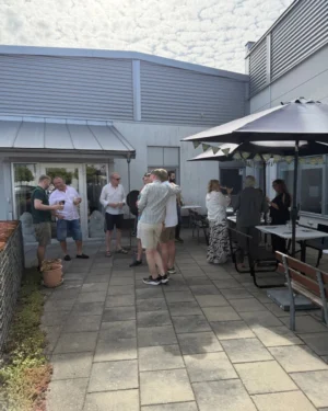 Grupp människor som minglar på en uteservering med plattor av sten. Några personer håller drycker, medan andra pratar med varandra. I bakgrunden syns ett fönster och en dörr som leder in i en byggnad, samt en stor parasoll med bänkar och bord. Det är en solig dag med en del moln på himlen.