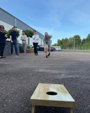 En grupp människor står på en asfalterad yta utanför en byggnad. En man i kortbyxor och randig skjorta kastar en liten väska mot en träplatta med ett hål i mitten, vilket är en del av ett spel. De andra i gruppen ser på, med varierande uttryck av intresse och uppmuntran. I bakgrunden syns bilar och gröna träd. Himmelns färg är ljusblå med några lätta moln.