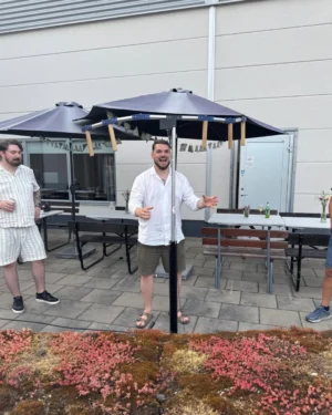 En man står under en svart parasoll med en glad uttryck och utsträckta armar, som om han talar till en grupp. Han har på sig en vit skjorta och shorts. I bakgrunden syns en annan man i randig klädsel som lyssnar. Det finns flera bord med stolar och några blommor på borden. Marken är täckt av en grön och rödaktig växtlighet.