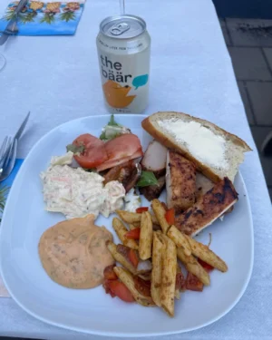 En vit tallrik med en uppdukad måltid som innehåller grillat kött, skivor av bröd med smör, en portion coleslaw, tomatskivor, en blandning av grönsallad, pasta med sås och bitar av paprika. En burk med läsk står bredvid på ett bord med ljusblå duk och en färgglad servett med ananasmönster.