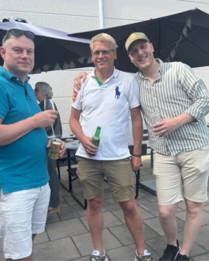 Tre män står tillsammans utomhus, leende och avslappnade. Den vänstra mannen har på sig en blå t-shirt och håller en burk i handen. Mannen i mitten bär en vit pikétröja med en grön och blå detalj samt ett par ljusa shorts och håller en grön flaska. Till höger står en man i en randig skjorta och ljusa shorts, som också håller i en burk. I bakgrunden syns parasoller och några andra personer.