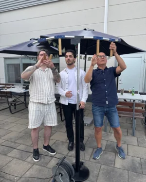 Tre män står under ett parasoll ute på en gård. Den första mannen till vänster har kort, mörkt hår, ett tatueringar på armen och bär en vit skjorta och rutiga shorts. Den andra mannen i mitten har mörkt hår och bär en vit skjorta över en svart T-shirt, med en drink i handen. Den tredje mannen till höger har kort, mörkt hår, glasögon och bär en mörk skjorta och jeansshorts. Alla tre verkar fokuserade på något som händer vid parasollet, där klädnypor är fästa. I bakgrunden syns ett bord med glas och flaskor.