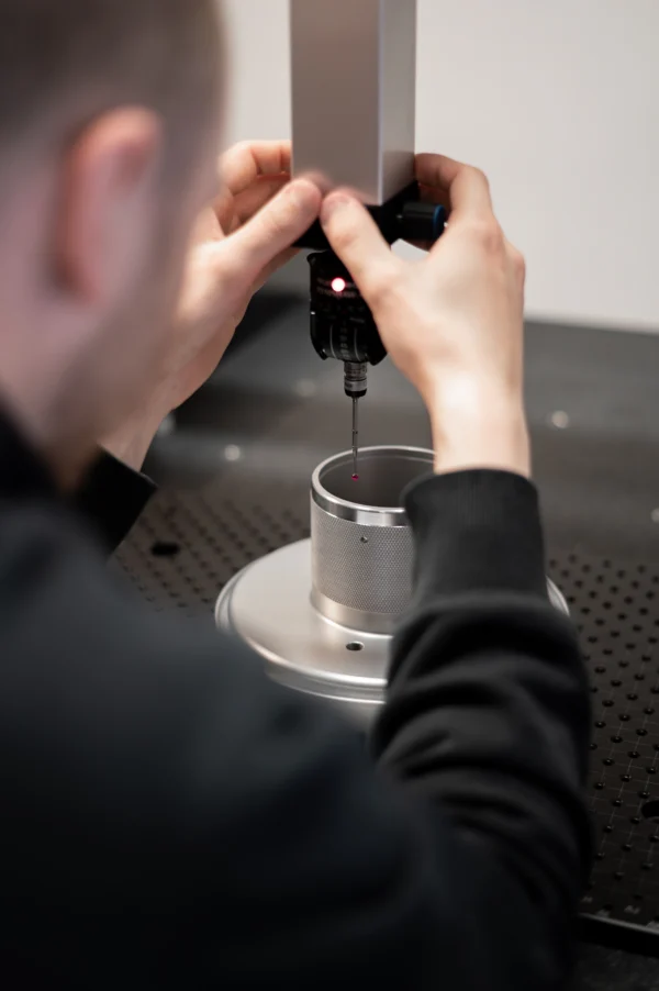 En person kontrollerar noggrant en cylindrisk metallkomponent med en mätinstrument med en röd ljuspunkt, som placeras ovanför komponenten på ett arbetsbord med ett mönster av små hål.