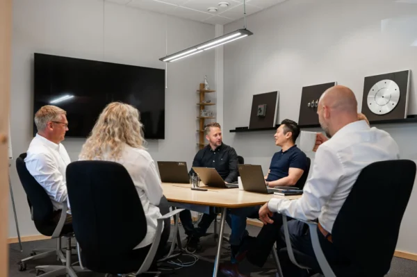 Mötesrum med fem personer som sitter runt ett bord. Två personer är vända mot varandra och diskuterar, medan tre andra sitter en bit bort och lyssnar. På bordet finns laptops och skrivmaterial. En stor TV-skärm hänger på väggen, och olika konstverk eller prover är uppsatta på en hylla i bakgrunden. Rummet är ljust och modernt inrett.